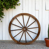 A large rustic wooden wagon wheel wall decor piece leaning against a white wood paneled wall on a gravel surface, with green foliage and a potted cactus nearby. 