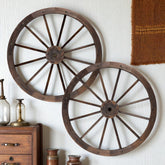 Two rustic wooden wagon wheels with dark brown finish and metal rivets, displayed on a white wall above a wooden cabinet with decorative glassware. 