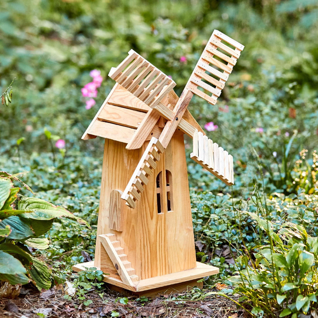 A small wooden garden windmill figurine with a natural light wood finish, set against a blurred green garden background. 