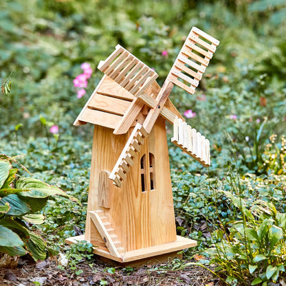 A small wooden garden windmill figurine with a natural light wood finish, set against a blurred green garden background. 