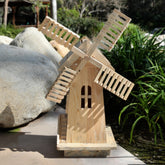 A small handcrafted wooden windmill garden ornament made from light natural wood, set outdoors on a stone surface with rocks and green plants in the background. 