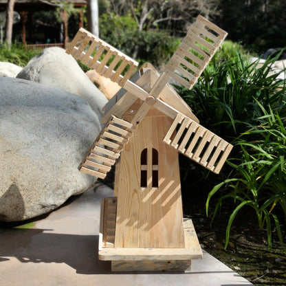 A small handcrafted wooden windmill garden ornament made from light natural wood, set outdoors on a stone surface with rocks and green plants in the background. 
