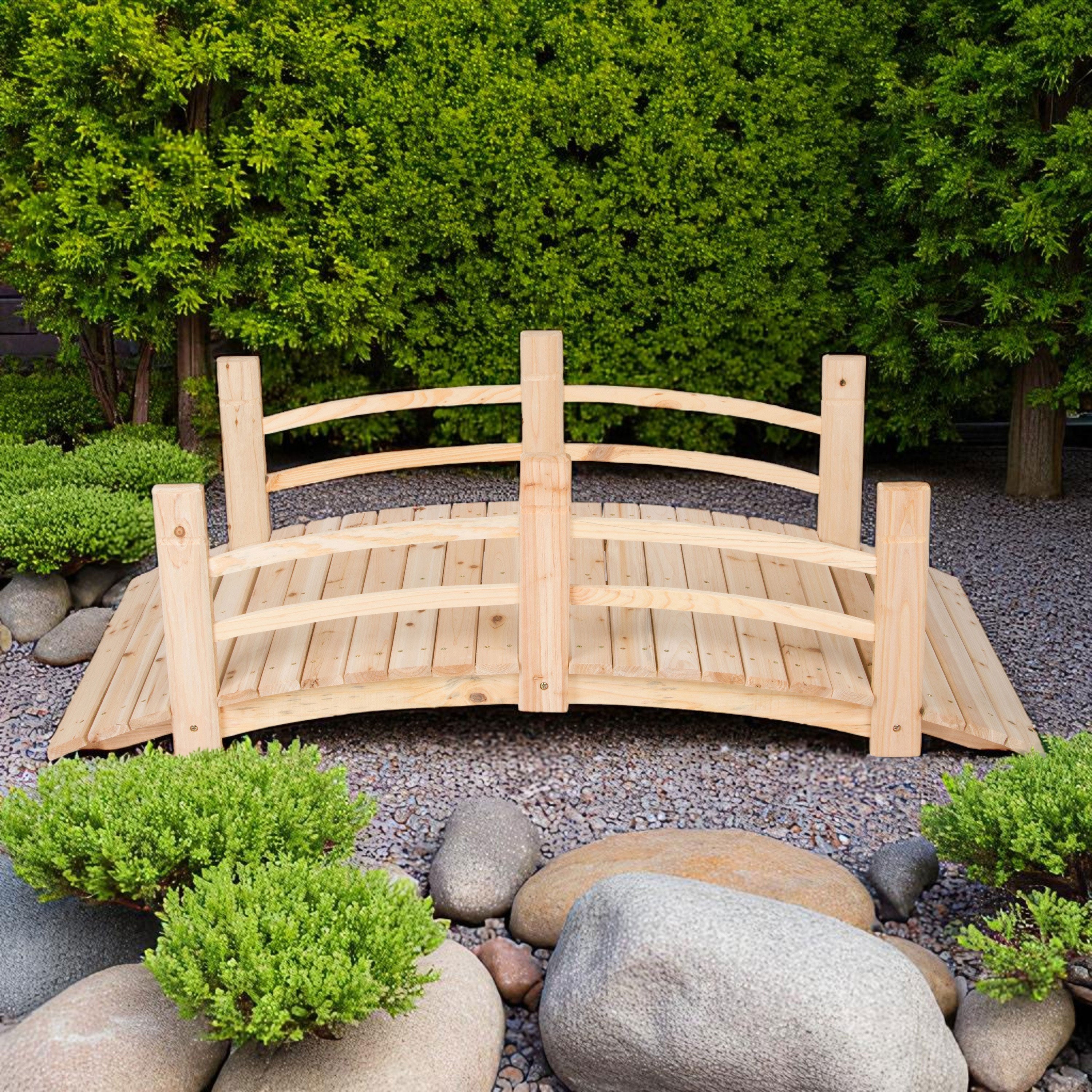 A small 5ft natural wood garden bridge with curved railings, shown in a landscaped yard with gravel, rocks, shrubs, and a green hedge background. 
