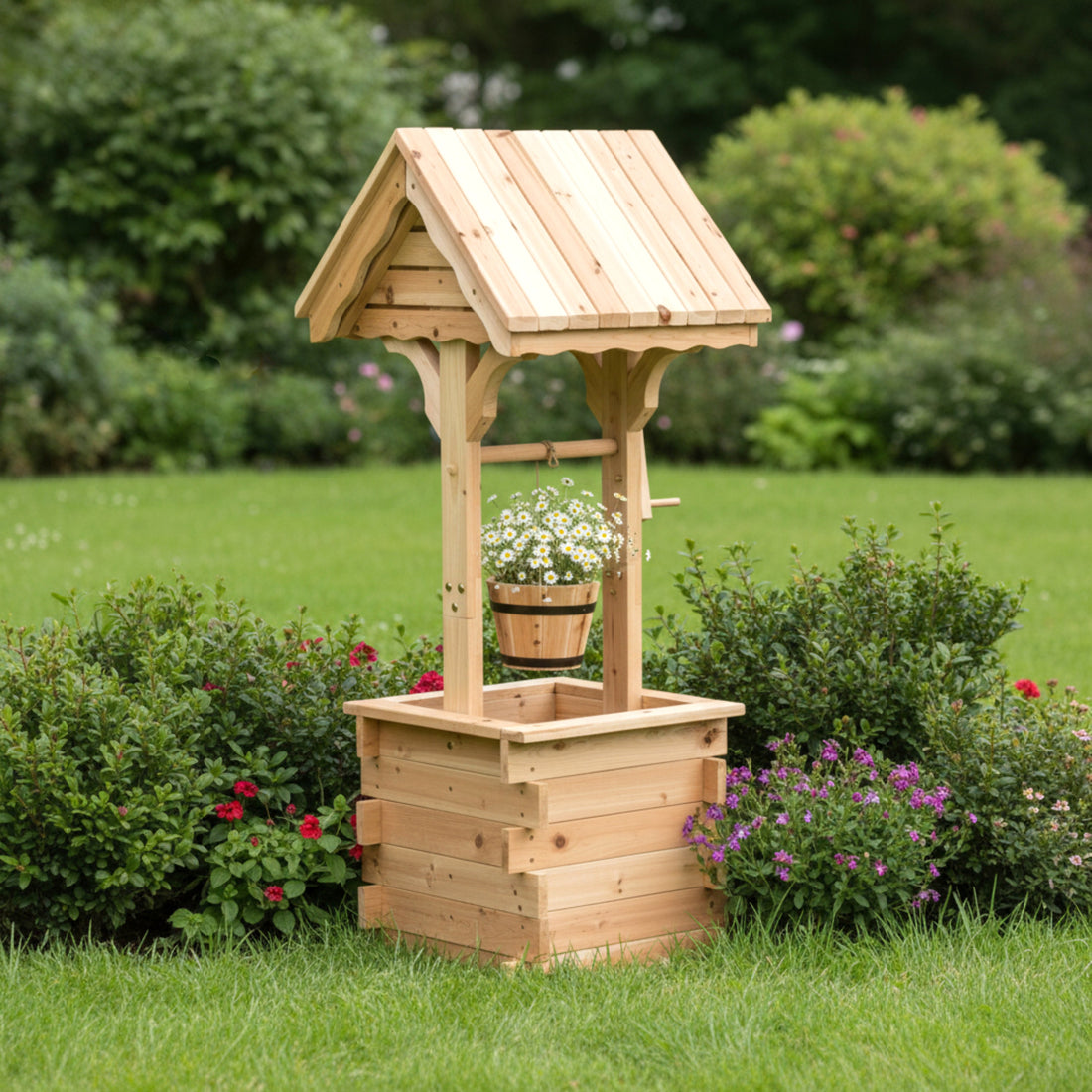 A small wooden garden well with a slatted bucket hanging from a curved roof, set on a grassy lawn surrounded by green shrubs and flowers. 