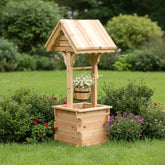 A small wooden garden well with a slatted bucket hanging from a curved roof, set on a grassy lawn surrounded by green shrubs and flowers. 