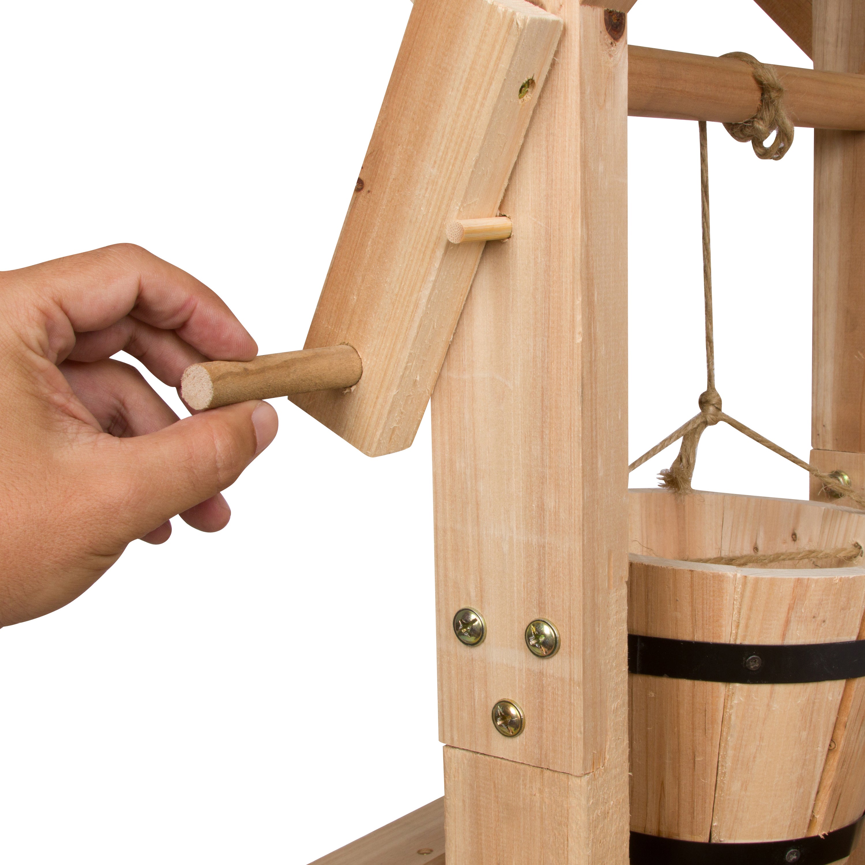 A close-up of a hand turning a wooden crank on a small wooden machine, with a bucket hanging on the right side against a white background. 