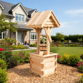 A light natural wood garden well structure with a slatted bucket and arched roof, placed on a wood chip bed in a landscaped backyard with a house and lawn in the background. 