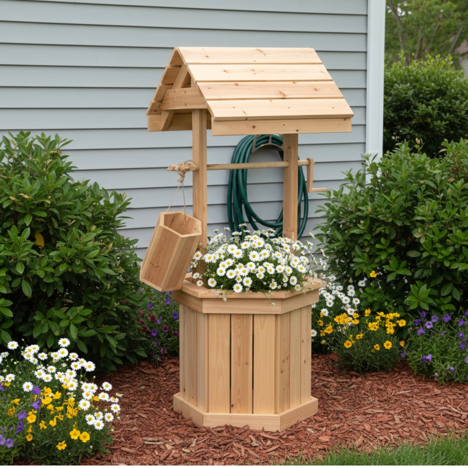 A wooden garden well planter and small well structure with a slatted bucket, shown outdoors in a landscaped garden bed in front of a gray house wall. 