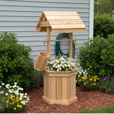 A wooden garden well planter and small well structure with a slatted bucket, shown outdoors in a landscaped garden bed in front of a gray house wall. 
