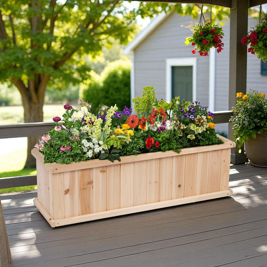 A large rectangular wooden planter box filled with colorful flowers, placed on a gray wooden deck in front of a house porch. Hanging flower baskets and another planter are visible in the background. 