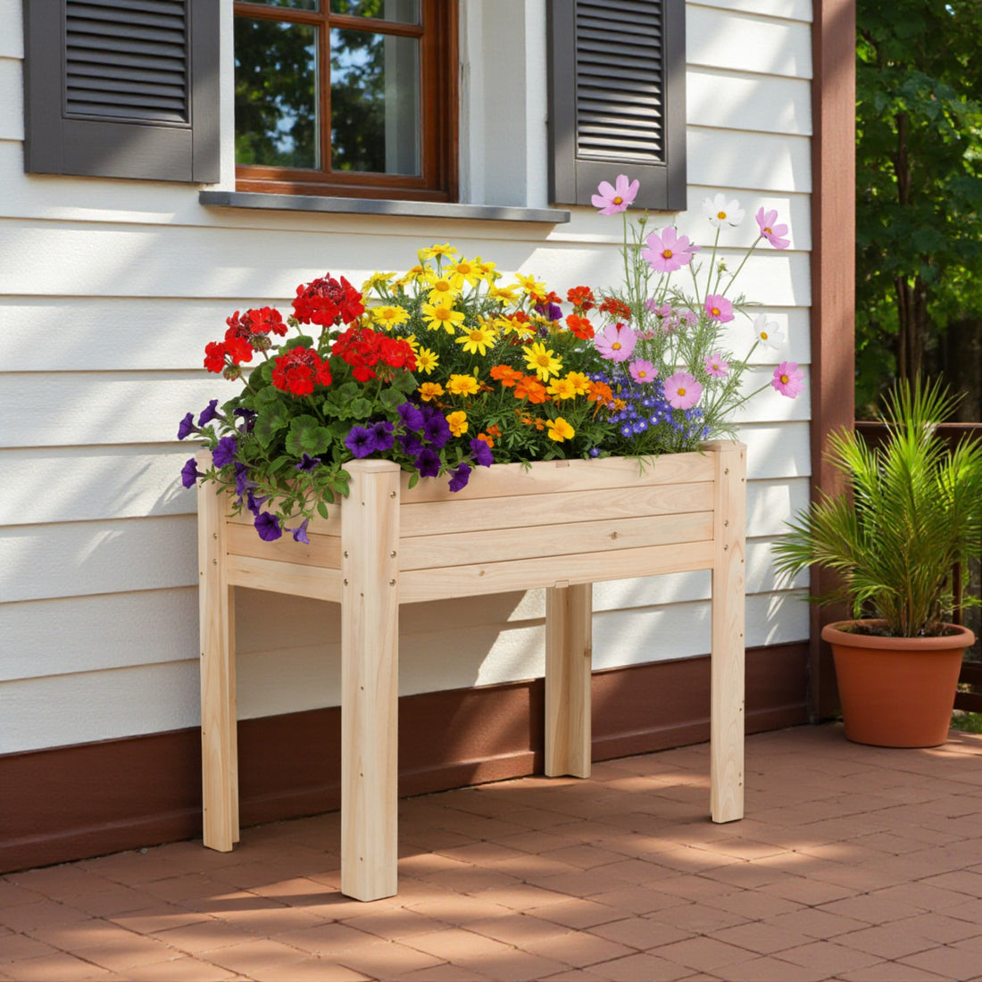 A light natural wood raised flower bed on a brick porch in front of a house with white siding and dark shutters, filled with colorful flowers including red, yellow, orange, purple, and pink blossoms. 