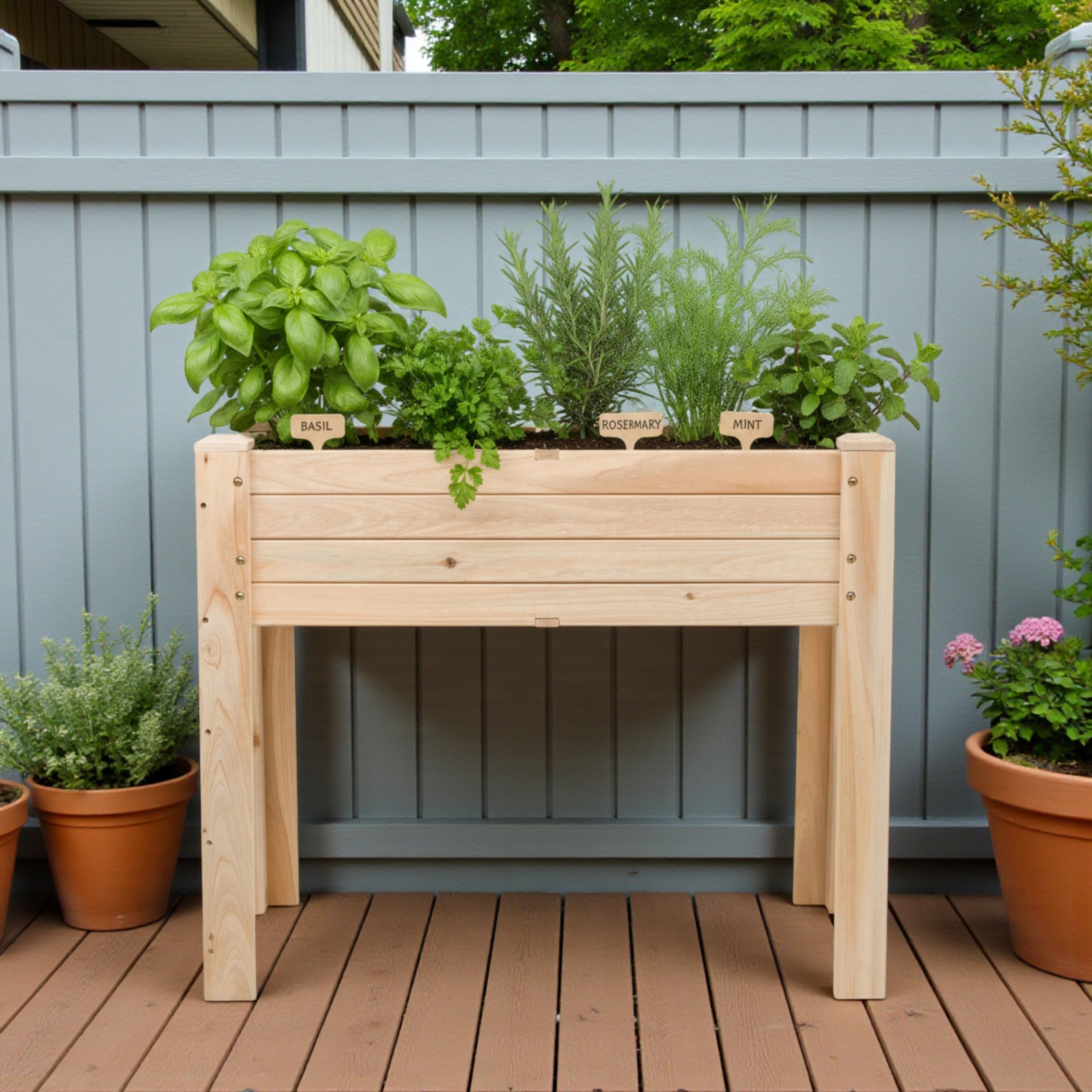A light natural wood raised garden bed planter table with four legs, shown on a brown wooden deck in front of a gray fence. The planter is filled with various green herbs, including basil, rosemary, and mint, with small wooden labels. 