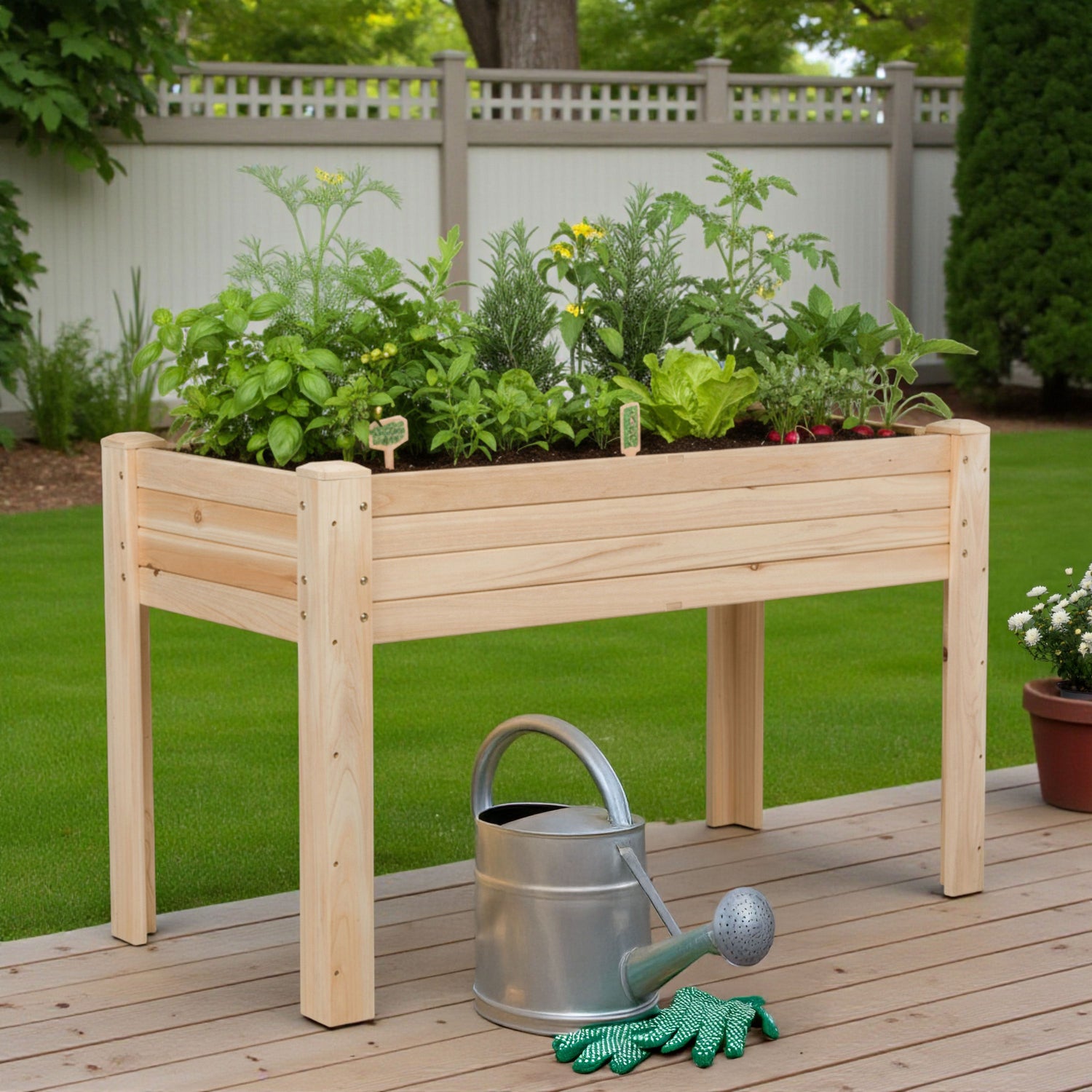 A light natural wood raised garden bed with a slatted frame, filled with various green plants, shown on a wooden deck in a backyard with a metal watering can and green gloves in front. 
