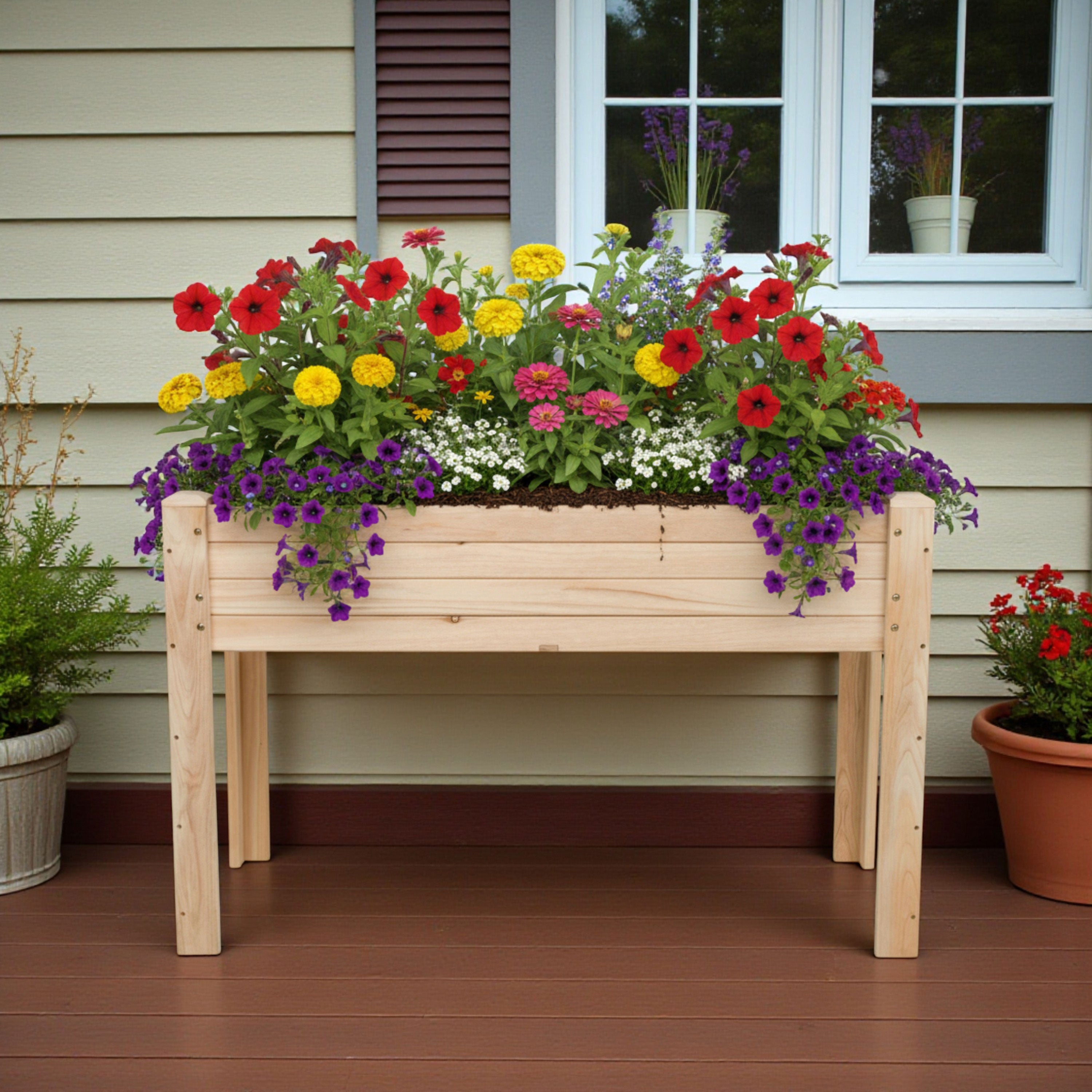 A rectangular wooden raised garden bed filled with colorful flowers, placed on a brown wooden deck in front of a house with beige siding and a window. 