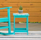 A turquoise painted wooden end table with a small potted cactus on top, set on a striped rug in front of a wooden fence. Part of a matching turquoise rocking chair is visible on the left. 