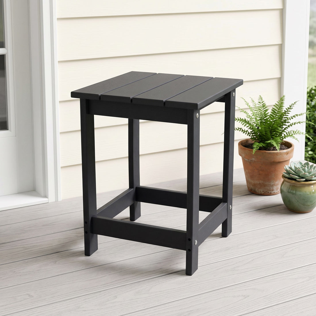 A black outdoor side table with a slatted top and open frame legs, shown on a light porch next to potted plants. 