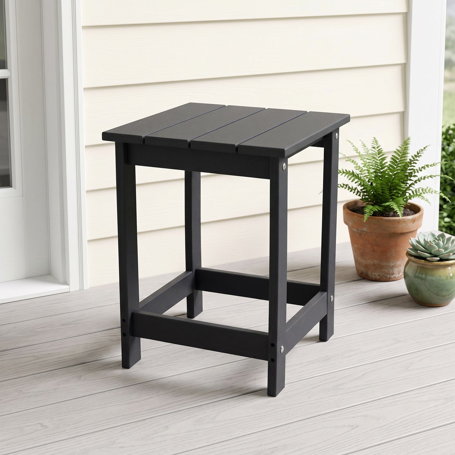 A black outdoor side table with a slatted top and open frame legs, shown on a light porch next to potted plants. 