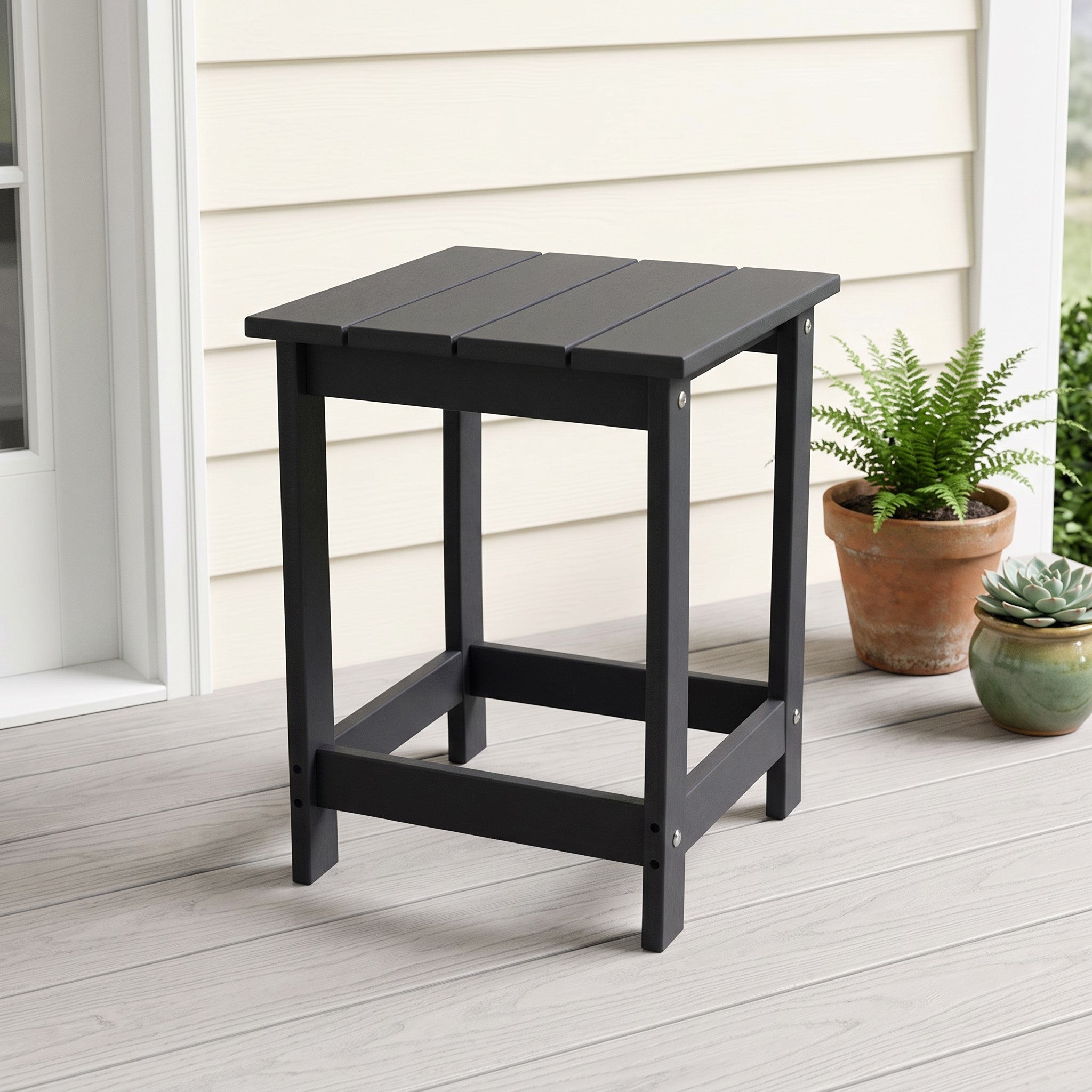A black outdoor side table with a slatted top and open frame legs, shown on a light porch next to potted plants. 