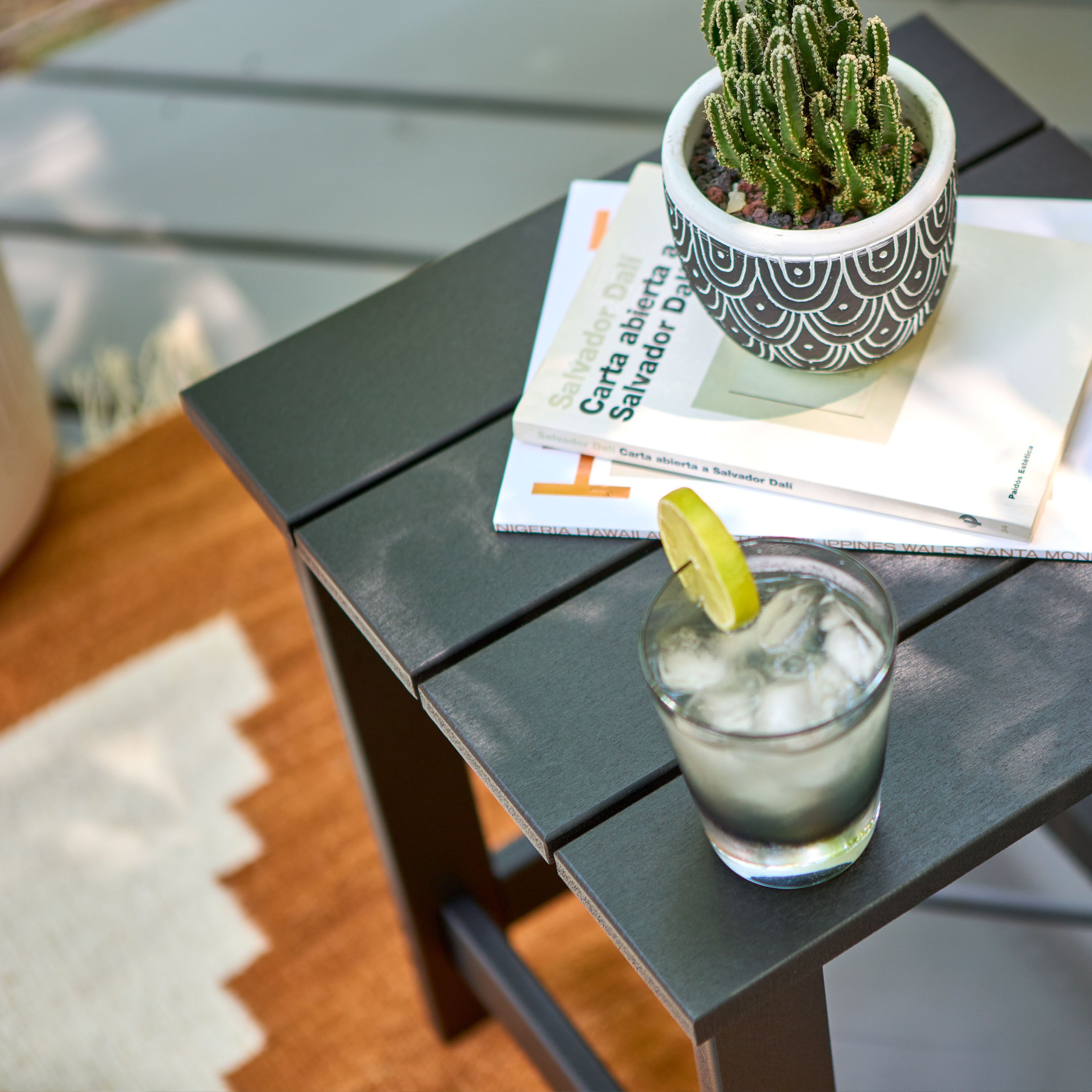 A black slatted outdoor side table in a patio setting, styled with a potted cactus, a magazine, and a glass of iced drink with a lime wedge. 