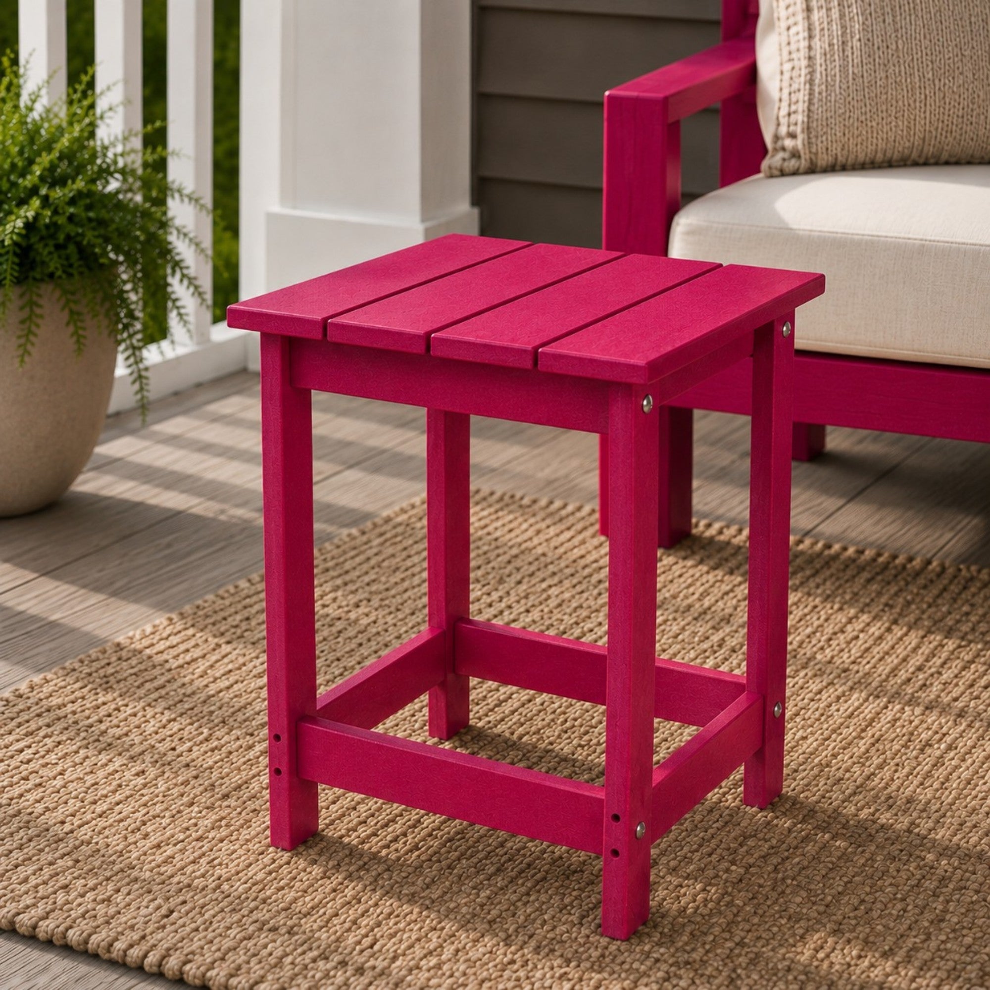 A red plastic side table with a slatted top and open lower shelf, shown on a woven rug in an outdoor patio setting next to a matching red chair. 