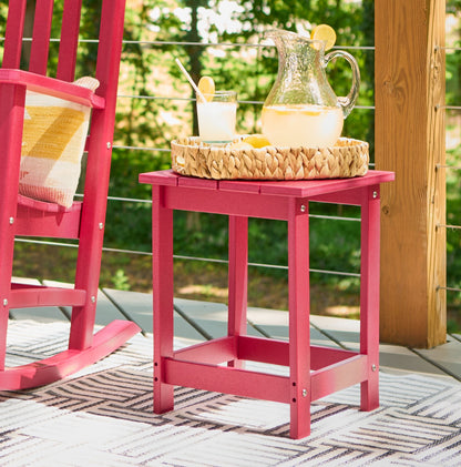 A red plastic side table on a patio, styled with a woven tray holding a glass pitcher of lemonade and a glass. A matching red rocking chair is partially visible on the left, with a green garden background. 