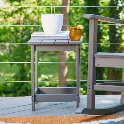 A gray outdoor side table with a slatted top and open lower shelf, styled on a deck with a white planter and amber drink on top. A matching gray rocking chair is partially visible at the right, with a blurred green garden background. 