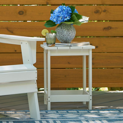 A white outdoor side table with a slatted top and open lower shelf, styled with a speckled vase of blue hydrangeas, a drink, and books, next to a white chair in front of a wooden fence. 