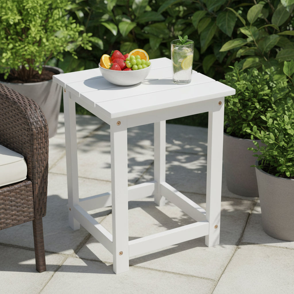 A white square outdoor side table on a patio, styled with a bowl of fruit and a glass of lemonade. The table is positioned next to a brown wicker chair, with green potted plants in the background. 