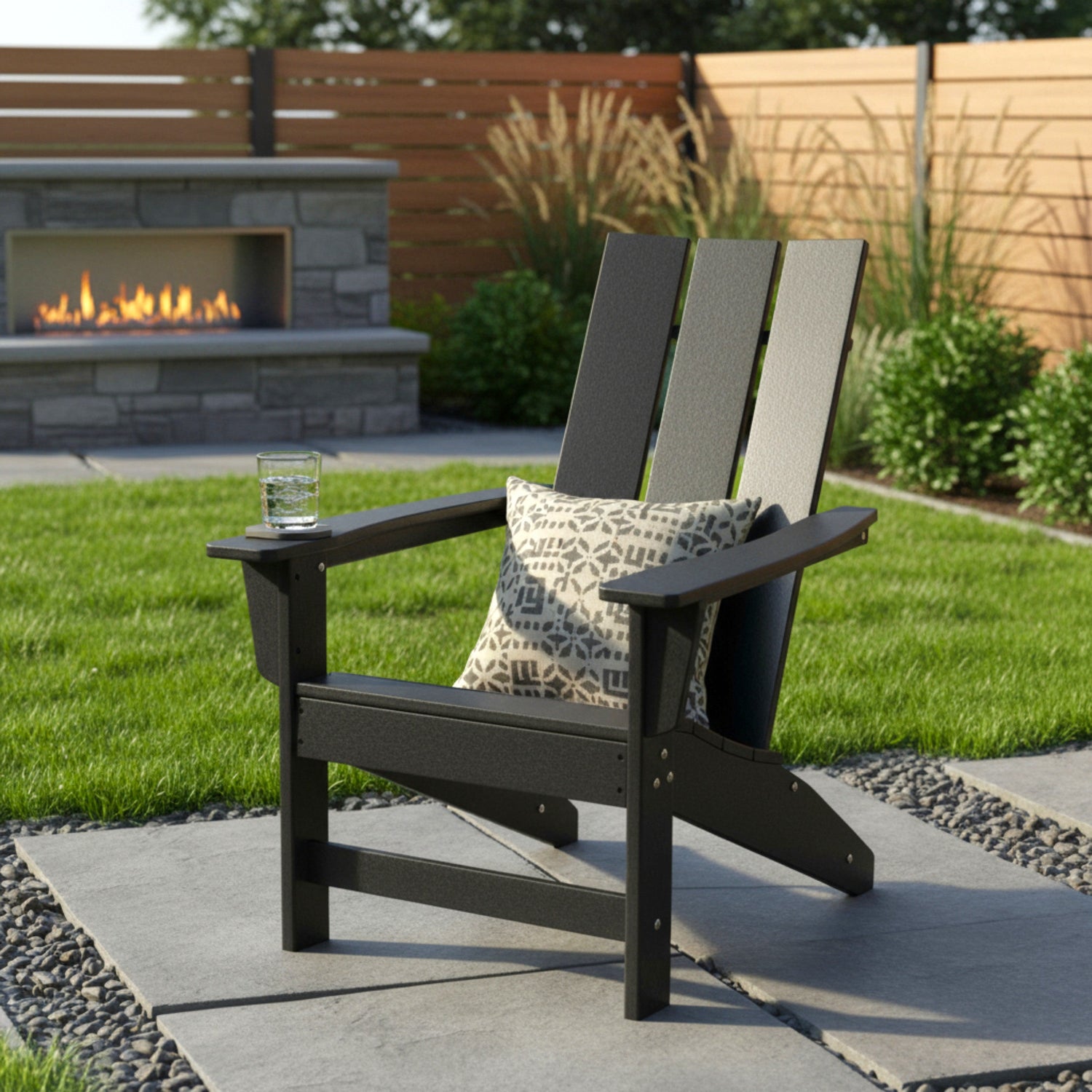 A black outdoor Adirondack chair on a stone patio with a patterned throw pillow and a glass on the armrest, set in a backyard with a green lawn, wooden fence, and a stone fireplace in the background. 