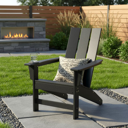 A black outdoor Adirondack chair on a stone patio with a patterned throw pillow and a glass on the armrest, set in a backyard with a green lawn, wooden fence, and a stone fireplace in the background. 