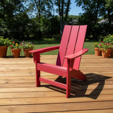 A bright red wooden Adirondack chair sits on a light brown wooden deck with a green lawn and trees in the background. 
