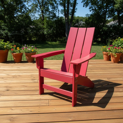 A bright red wooden Adirondack chair sits on a light brown wooden deck with a green lawn and trees in the background. 