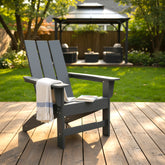 A dark gray outdoor Adirondack chair sits on a wooden deck in a backyard setting, with a white knit throw blanket draped over the arm and a book resting on the seat. A green lawn, trees, and a black patio gazebo are visible in the background. 