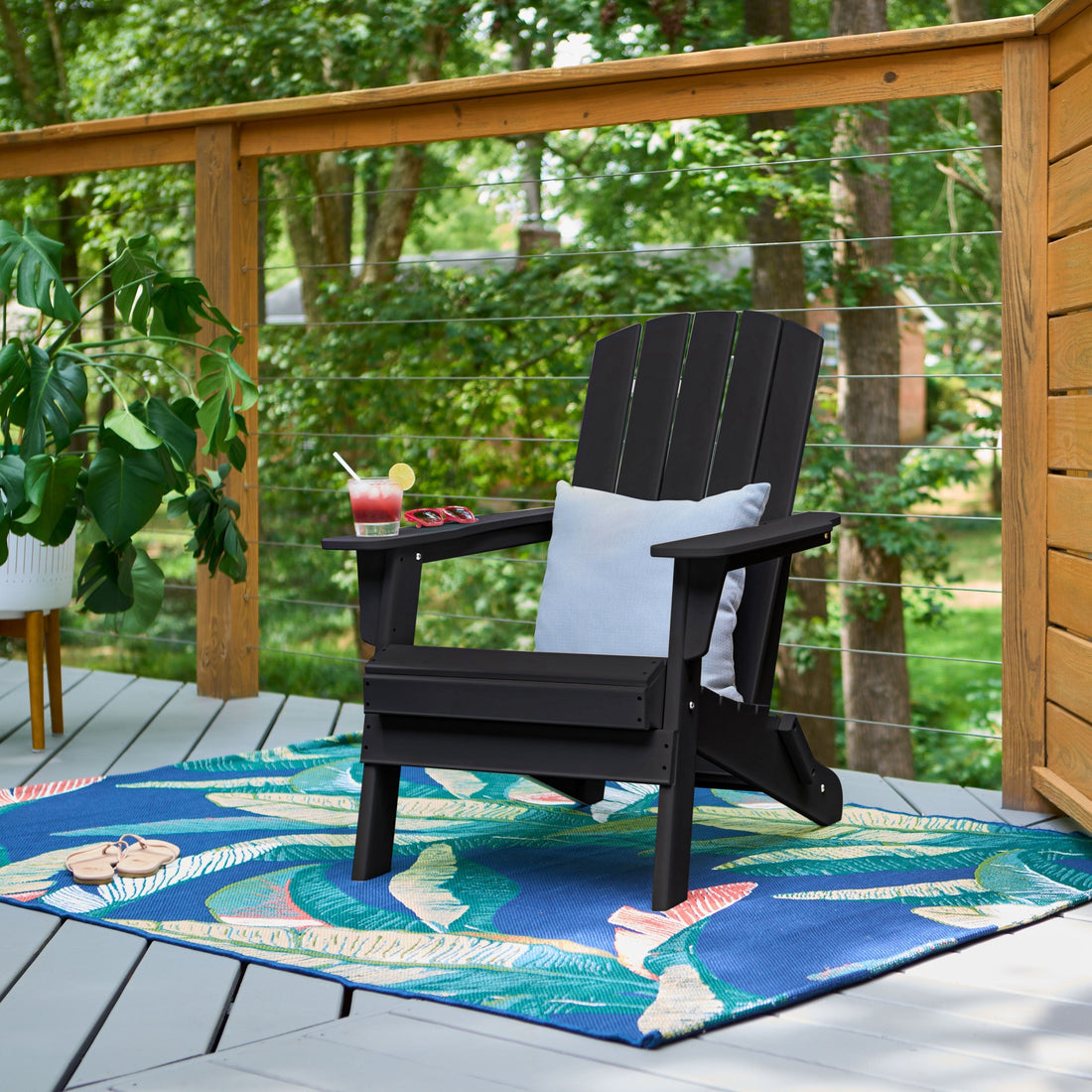 A black outdoor Adirondack chair with a white pillow sits on a blue tropical-patterned patio rug on a wooden deck, with a drink on the armrest and a wooden railing with green trees in the background. 