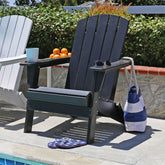 A black Adirondack chair sits on a patio poolside with a blue and white striped tote bag hanging from its arm, sunglasses on the armrest, and a pair of blue-and-white flip-flops on the seat. Another white chair is partially visible on the left, with green foliage and a stone wall in the background. 