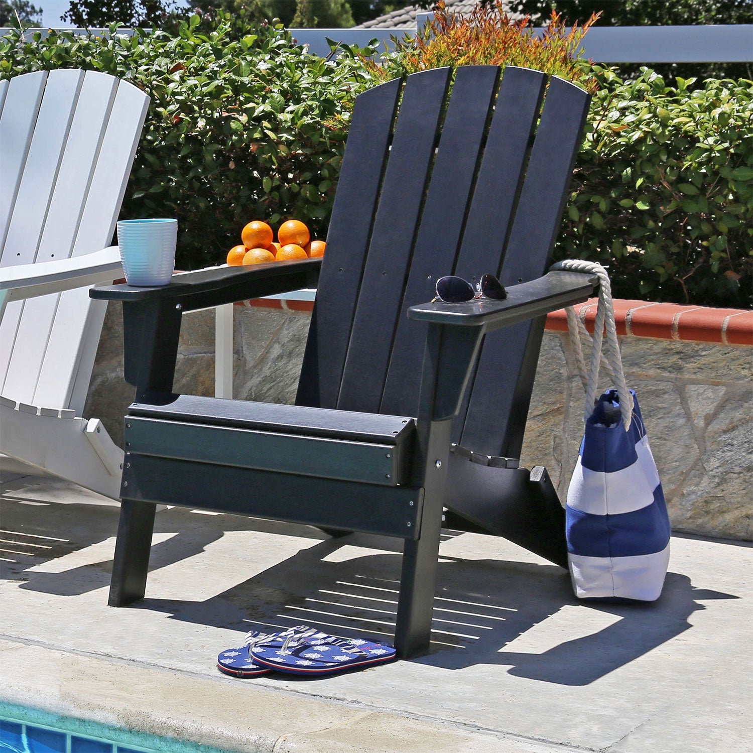 A black Adirondack chair sits on a patio poolside with a blue and white striped tote bag hanging from its arm, sunglasses on the armrest, and a pair of blue-and-white flip-flops on the seat. Another white chair is partially visible on the left, with green foliage and a stone wall in the background. 