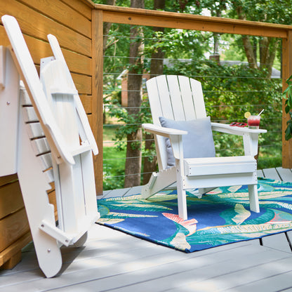 A white Adirondack-style outdoor chair sits on a patterned blue tropical rug on a wooden deck, with another folded chair leaning against the wall and a drink on the armrest. Trees and a railing are visible in the background. 
