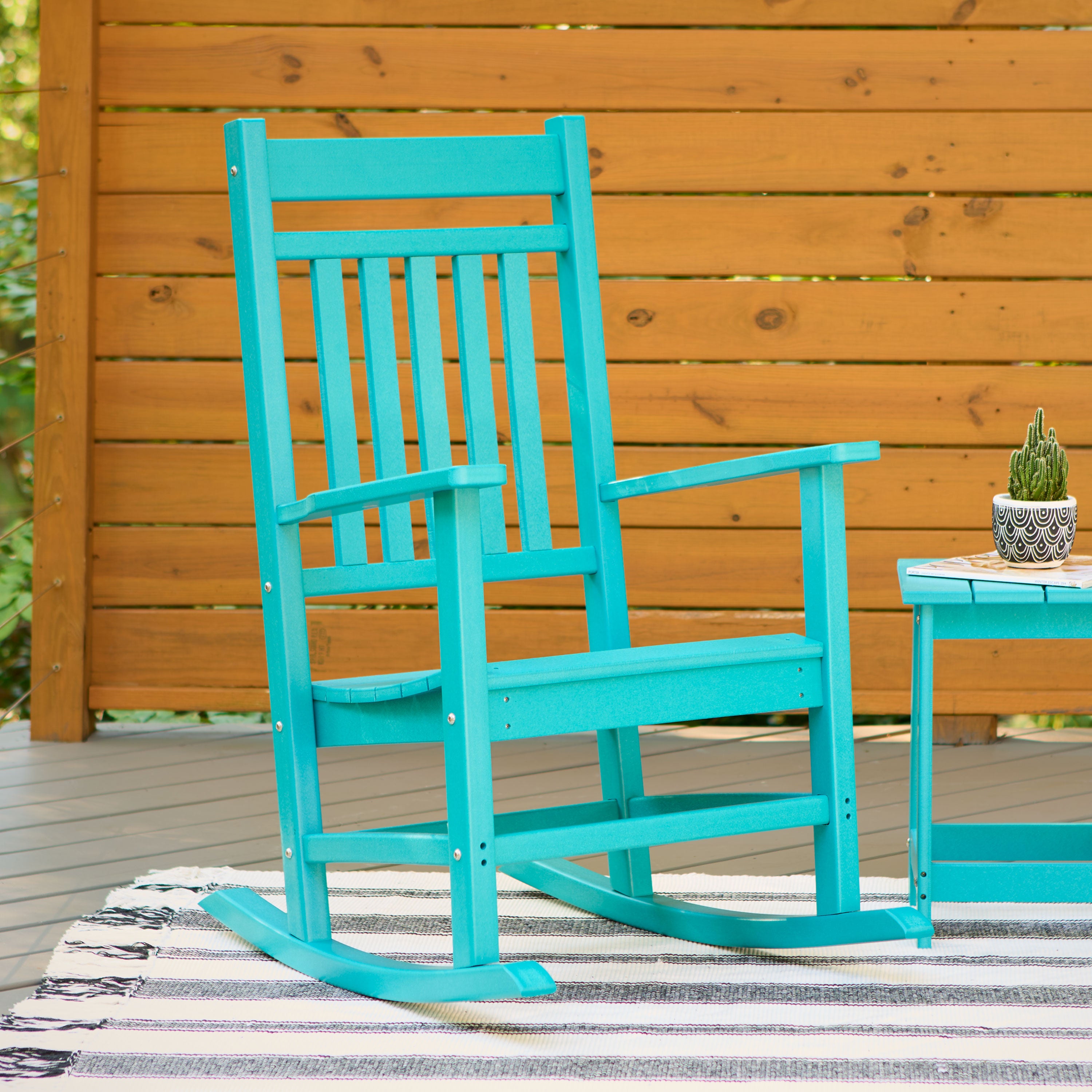 A turquoise wooden rocking chair with a matching side table, set on a striped rug in front of a wooden fence. 