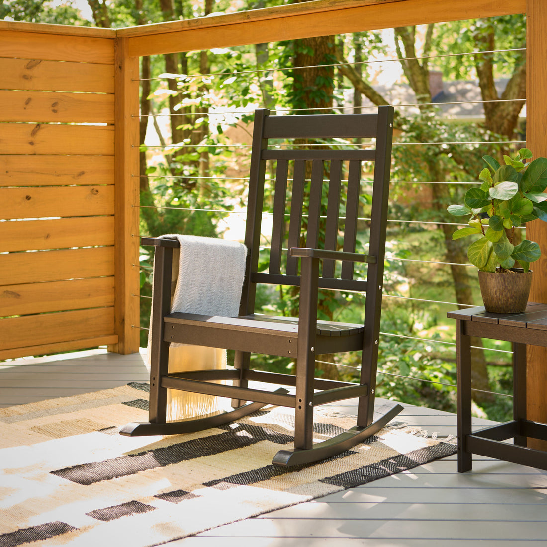 A black plastic rocking chair with a white throw blanket sits on a patterned rug on a wooden deck, with a side table and a potted plant next to it. The background shows a lush green garden visible through a wooden railing. 