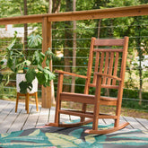 A brown plastic rocking chair on a deck with a tropical leaf-patterned rug, a potted plant on a small stool, and a forest background visible through a cable railing. 