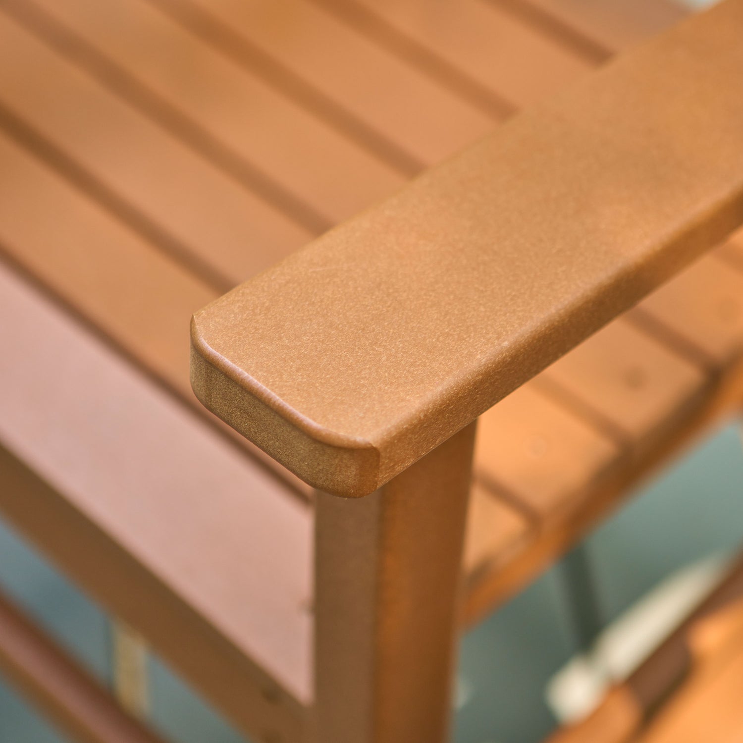 Close-up of a brown slatted outdoor chair or bench with a rounded armrest corner, showing the textured recycled plastic finish and slatted backrest.  