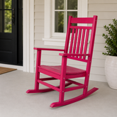 A red recycled plastic rocking chair with a slatted back and armrests, positioned on a light gray concrete porch in front of a white-paneled house with a black door. 