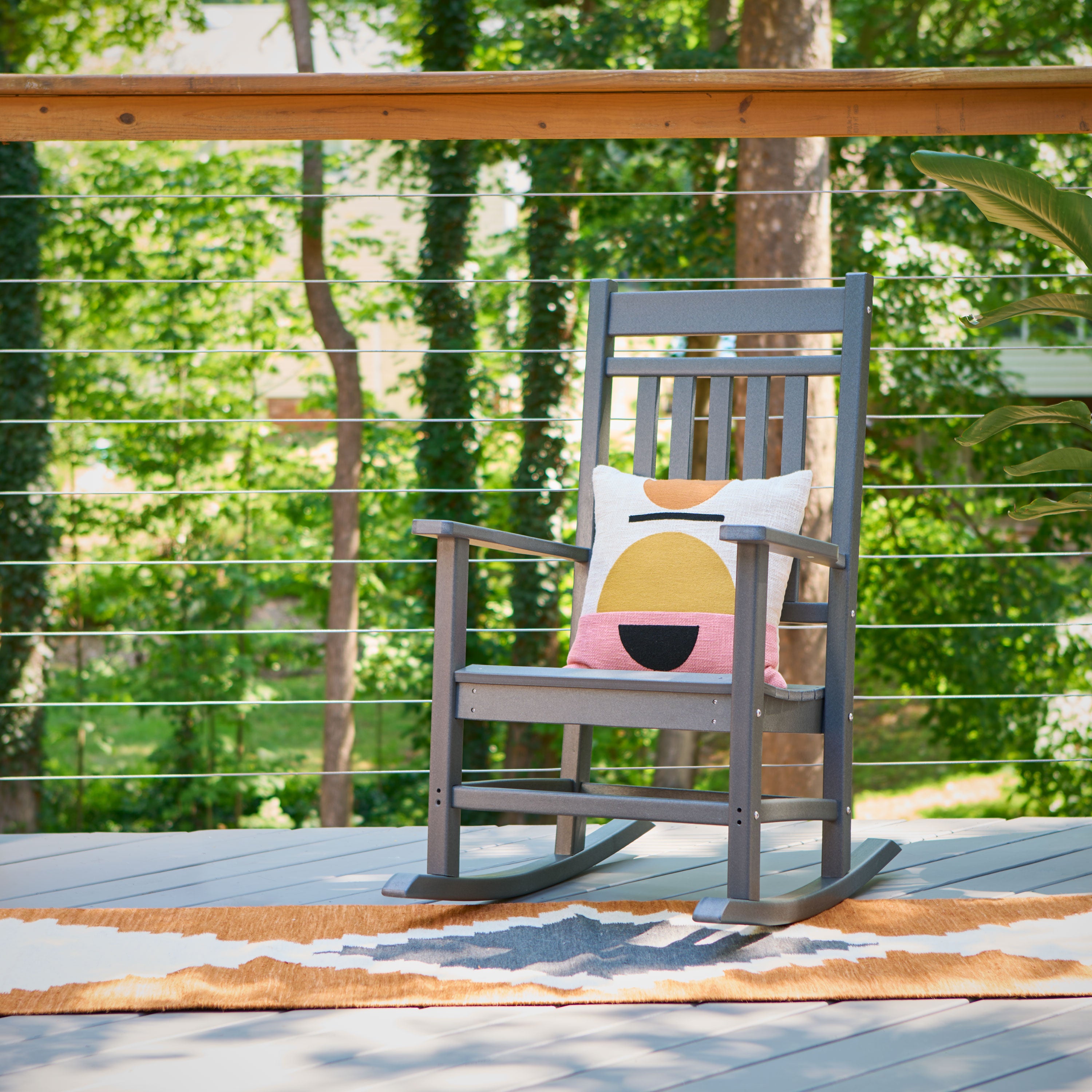 A gray wooden rocking chair sits on a deck with a colorful geometric throw pillow, set against a blurred background of green trees and a wooden railing. 