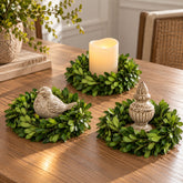 Three real green boxwood wreath table decor pieces arranged on a wooden dining table, including a wreath with a stone bird, a wreath with a lit pillar candle, and a wreath with an ornate urn-shaped accent.
