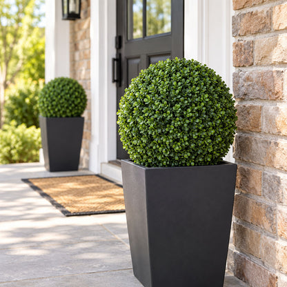 A large black square planter holding a dense, rounded green boxwood topiary, placed on a stone patio beside a brick house exterior with a matching second planter in the background.