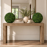 A rustic wooden console table styled in a bright, modern entryway. The table is decorated with two spherical green topiary trees, a textured beige ceramic vase with olive branches, a small white candle, and a stack of books.