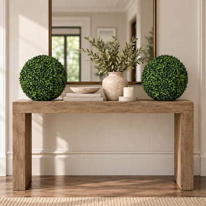 A rustic wooden console table styled in a bright, modern entryway. The table is decorated with two spherical green topiary trees, a textured beige ceramic vase with olive branches, a small white candle, and a stack of books.